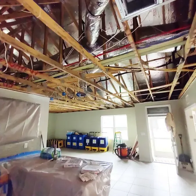 An open ceiling reveals framing and ducts above protected furniture, equipment and a bright doorway.
