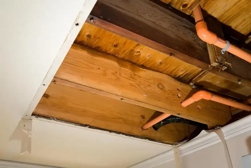 An opened ceiling reveals wooden joists and orange pipes above a tiled wall.