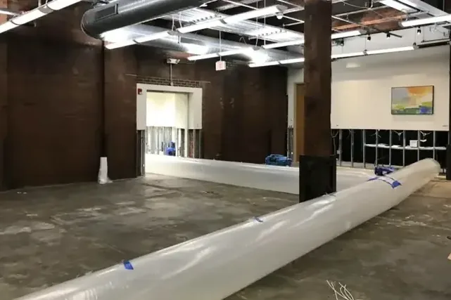 A long white drying duct crosses an open commercial room beneath exposed ceiling services.