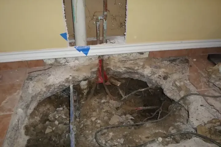 An opening in a concrete floor exposes pipes beside an opened wall.